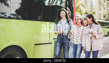 Fröhliche asiatische Freunde, die Smartphones am Busbahnhof nutzen - Junge Studenten, die nach der Schule im Freien Spaß haben - Freundschaft, Techniktrends, Universität und Stockfoto