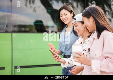 Fröhliche asiatische Freunde, die Smartphones am Busbahnhof nutzen - Junge Studenten, die nach der Schule Spaß mit der Phones App haben - Freundschaft, Universität und Tras Stockfoto