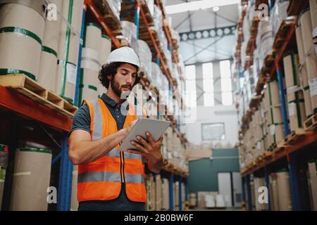 Der junge kaukasische Ingenieur in Hardhat einen Tablet-Computer in einer Schwerindustrie verwendet, der aus Sicherheitsgründen einen weißen Helm und eine orangefarbene Weste trägt Stockfoto