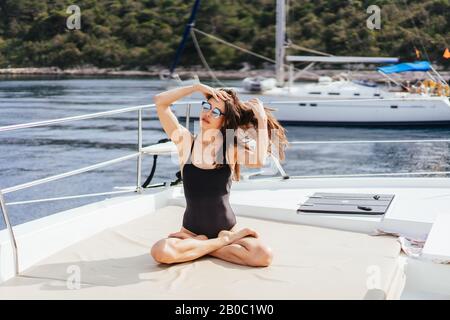 Junge gesunde und ruhige Frau, die auf dem Segelyacht-Boot im Meer auf Inselhintergrund Yoga macht Stockfoto