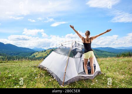 Rückblick auf den Touristenwanderweg der jungen Frau, der im Zelt steht, die Hände in die Luft hebt und den sonnigen Sommermorgen in den Bergen genießt. Abenteuer Urlaub Outdoor-Konzept Stockfoto