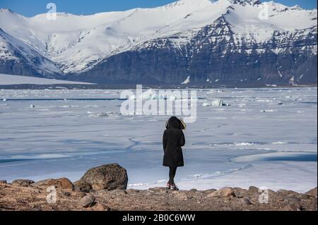 Rückansicht der jungen Frau auf Felsvorsprung mit Blick auf den halbgefrorenen Gletscher, Jokulsarlon, Island. Stockfoto