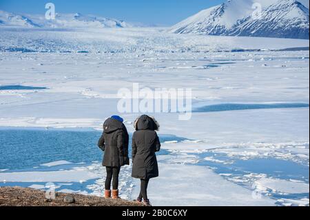 Rückansicht von zwei jungen Frauen, die auf Felsvorsprung stehen und die Aussicht auf den halbgefrorenen Gletscher Jokulsarlon, Island genießen. Stockfoto