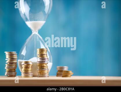 Uhr mit gestapelten Münzen und Sanduhr auf blauem Hintergrund Stockfoto