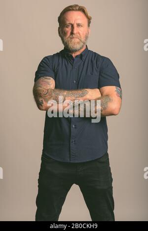 Studio shot reifer bärtiger Mann mit hand Tattoos stehend mit Waffen in grauer Hintergrund gekreuzt Stockfoto