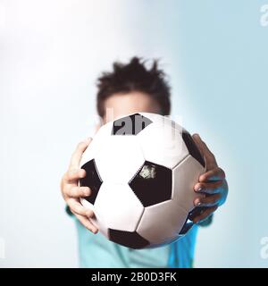 Ein Junge in ein blaues T-Shirt mit einem Fußball in der Hand seinen Kopf verdeckt auf einem blauen Hintergrund Stockfoto