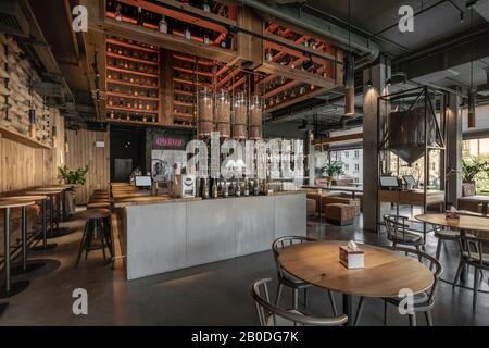 Stilvolles Kaffeehaus im Loft-Stil mit großen Panoramafenstern und Betonsäulen. Es gibt Holztische mit Sofas und Stühlen, Regale mit Flasche Stockfoto