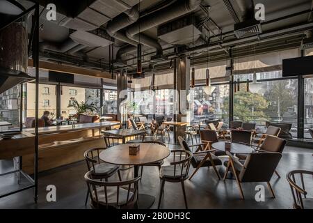 Schönes Kaffeehaus im Loft-Stil mit großen Panoramafenstern und Betonsäulen. Es gibt Holztische mit Sofas und Stühlen und Sesseln, g Stockfoto