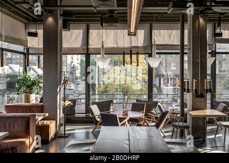 Trendige Kaffeehaus im Loft-Stil mit großen Panoramafenstern und Betonsäulen. Es gibt Holztische mit Sofas und Stühlen und Sesseln, Glas Stockfoto