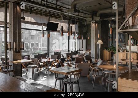 Schönes Kaffeehaus im Loft-Stil mit großen Panoramafenstern und Betonsäulen. Es gibt Holztische mit Stühlen und Sesseln, eine gläserige Kaffeebohne Stockfoto