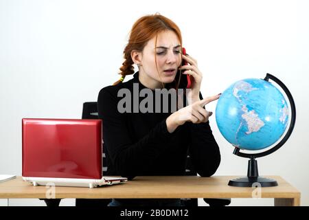 Junge Reisebüro-Frau hinter dem Schreibtisch mit Laptop und geografischer Weltöffentlichkeit, die auf einem Handy spricht. Stockfoto