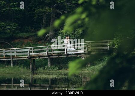 Der Bräutigam mit der Braut oder ein Mann mit einer Frau auf einer Brücke in einem Wald in der Nähe von einem See. Hochzeit in der Natur oder Einsamkeit für Zwei. Stockfoto