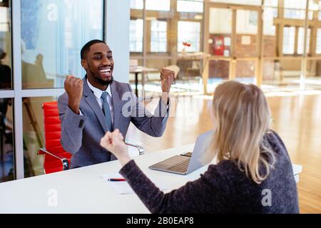 Porträt eines fröhlichen lächelnden jungen Mannes im Business-Anzug, der mit Fäusten gefeiert wird, mit einer Frau, einem Kollegen oder einem Kunden im Bürotreffen Stockfoto