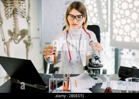 Junge hübsche kaukasische Wissenschaftlerin, die sich die wissenschaftliche Probe ansieht, Reagenzglas mit roter Flüssigkeit in der Hand, während sie mit Laptop am Tisch sitzt Stockfoto