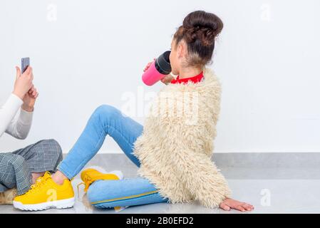 Portrait des niedlichen Teenager-Modells, das heißen Kaffee aus wiederverwendbarer pinkfarbener Tasse trinkt und ihre Freundin posiert. Stockfoto