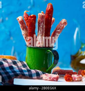 Einige Fuet- und Chorizo-Stücke, zwei verschiedene spanische Wurstwaren in einem grünen Becher auf einem Tisch, neben einigen Brotscheiben und einem Grausamkeiten mit Oliven Stockfoto