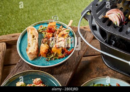 Gegrillte Gemüsekebabs mit Tofu, Kürbis, Pilzen und Tomaten auf einem Teller mit geröstetem Brot auf einem Gartentisch neben einem tragbaren Grill Stockfoto
