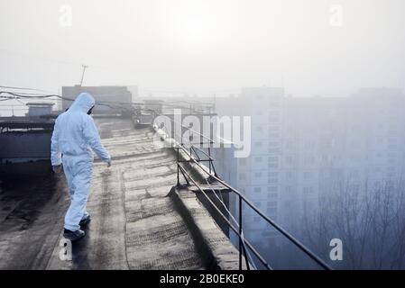 Rückansicht des Mannes, der weißen Overall und Gasmaske trägt und auf dem Dach eines städtischen Gebäudes spaziert. Vor dem Hintergrund der aufgehenden Sonne bei nebeligem Wetter, Konzept des Umweltproblems Stockfoto