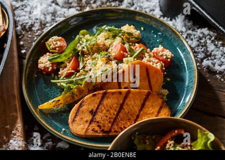 Gegrillte Süßkartoffelscheiben mit Quinoa oder Couscousalat mit Tomaten und frisch grüner Rakete auf einem Tisch im Freien mit Winterschnee in der Nähe Stockfoto