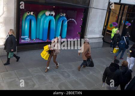 Mit drei gelben Einkaufstaschen geht eine Dame in das Kaufhaus Selfridges an der Londoner Oxford Street und passiert eine themenbezogene Fensteranzeige, die einige Cartoon-Charaktere enthält, die der Öffentlichkeit am 7. Februar 2020 ein glückliches neues Jahrzehnt wünschen, in London, England. Stockfoto