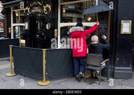 Geschützt von einem Regenschirm, der über ihrem Kopf gehalten wird, malt eine Frau, die Künstlerin, die signautarke, den Text des Essensmenüs eines West End Pubs mit Pies und Fish & Chips vor Dem Porcupine, einem traditionellen britischen Pub an der Charing Cross Road, am 17. Februar 2020, in London, England. Stockfoto