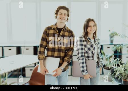 Junger Mann und Frau, die Laptops im Arbeitszimmer halten und lächeln. Mädchen hält Notizbuch in der Hand Stockfoto