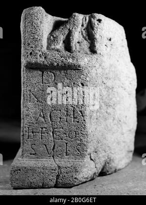 Linker unterer Teil eines Altars mit Nische. Kalksteine. Links in der Nische steht ein Hund, der bis zur Mitte gewöhnt ist. Der Kopf ist verschwunden. In dieser Höhe wurde der gesamte obere Teil des Altars abgebrochen, so ein Bruch, der bereits in der Antike bestand, und man bezeugen die Anwesenheit von zwei großen Eisenheften, einer links und einer hinten. Rechts neben dem Hund sieht man die Falten einer Robe. Von dort aus ist der Flügelaltar schräg nach hinten abgeschnitten. Nur an der Vorderseite befindet sich eine stark hervorstehende Liste zwischen der Aufschrift und der Nische. Vorne und seitlich ist eine Sockelplatte angebracht. Auf der linken Seite befindet sich eine Anlage Stockfoto