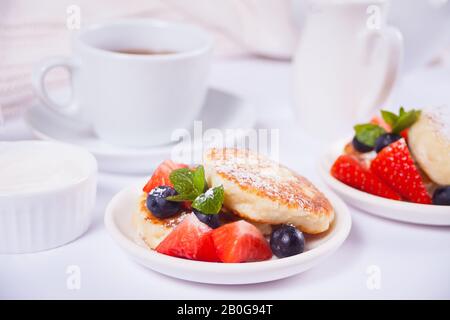 Zwei Teller mit Hüttenkäse pfanniki syrniki mit Beeren und Puderzucker. Tasse Tee, Teekanne und Milchkanne im Hintergrund Stockfoto