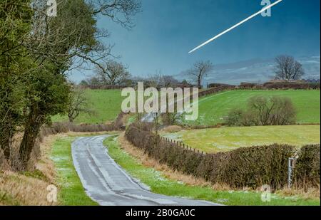 Icy Road mit leicht bedeckten Schneehügeln in der Ferne. Ein Düsenflugzeug fährt zum Flughafen Glasgow und verlässt einen Dampfpfad über den Himmel. Stockfoto