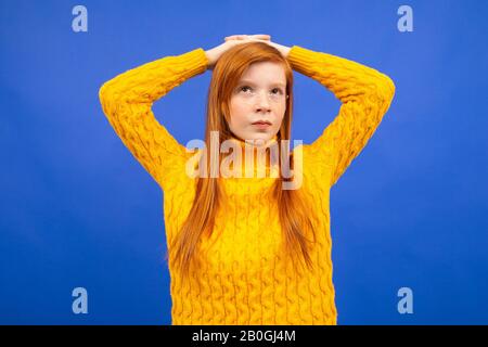 Europäisches rothaariges Teenager-Mädchen in einem gelben Pullover steht nachdenklich und hält seine Hände hinter dem Kopf auf einem blauen Studiohintergrund Stockfoto