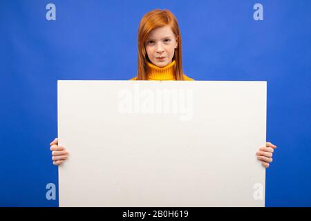 Charmantes europäisches rothaariges Teenager-Mädchen, das ein leeres Papierposter mit einem Mockup für Werbung auf blauem Studiohintergrund hält Stockfoto