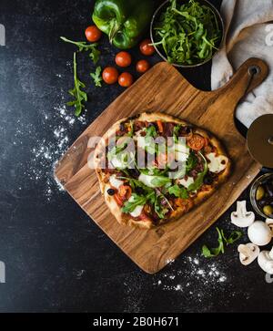 Draufsicht auf handgemachte Pizza auf Holzbrett mit Zutaten um sie herum. Stockfoto