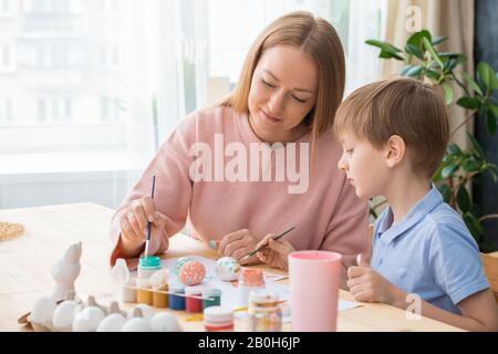 Attraktive junge Mutter und Sohn malen während der Ostervorbereitung Eier in leuchtenden Farben Stockfoto