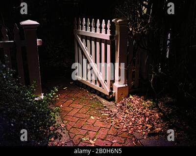 Tor des weißen Pfostenzauns öffnet sich in der Dunkelheit, Backsteinpfad. Stockfoto