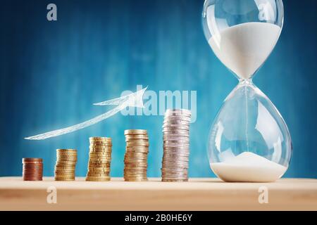 Uhr mit gestapelten Münzen und Sanduhr auf blauem Hintergrund Stockfoto