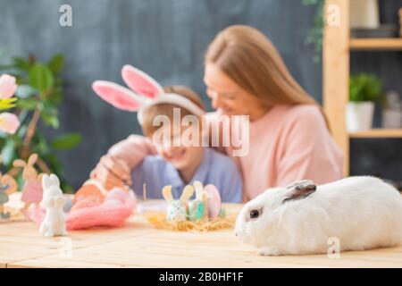 Nahaufnahme des weißen, flauschigen Kaninchens, das auf Holztisch mit Osterdekorationen liegt, Mutter umarmt den Sohn im Hintergrund Stockfoto