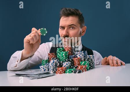 Mann in schwarzer Weste und Hemd am weißen Tisch sitzend mit Stapel Chips und Bargeld darauf, posiert auf blauem Hintergrund. Glücksspiel, Poker, Casino. Nahaufnahme. Stockfoto