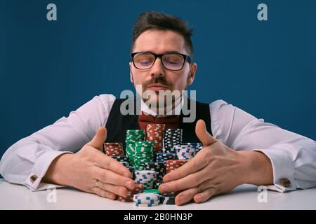 Mann in Brille, schwarze Weste und Hemd am weißen Tisch mit darauf aufsitzenden Spitenstapeln auf blauem Hintergrund. Glücksspiel, Poker, Casino. Nahaufnahme. Stockfoto