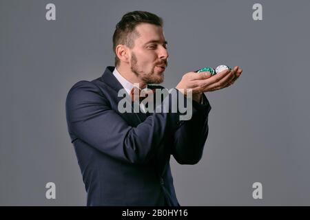 Mann in schwarzem klassischem Anzug und roter Krawatte mit einigen farbigen Chips, posiert vor grauem Studiohintergrund. Glücksspiel, Poker, Casino. Nahaufnahme. Stockfoto