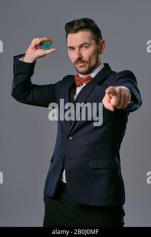 Mann in schwarzem klassischem Anzug und roter Krawatte mit einigen farbigen Chips, posiert vor grauem Studiohintergrund. Glücksspiel, Poker, Casino. Nahaufnahme. Stockfoto