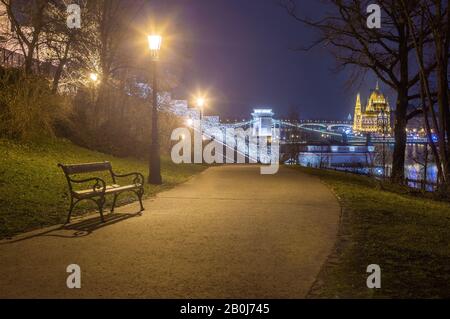 Budapest, Ungarn - Bank- und Lampenposten in einem Park im Buda-Viertel mit Szechenyi-Kettenbrücke und Parlament im Hintergrund zur Winterzeit Stockfoto