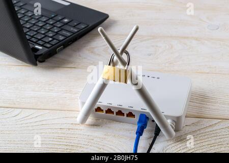 Weißer WLAN-Router mit Vorhängeschloss auf Antennen in der Nähe des Laptops über Holztisch. Verbotene oder eingeschränkte Konzepte der Internet- und Internetzensur. Stockfoto