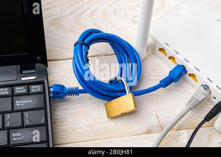 Vorhängeschloss auf einem Netzwerkkabel, das Laptop und weißen Wi-Fi-Router auf einem Holztisch verbindet. Verbotene oder eingeschränkte Konzepte der Internet- und Internetzensur Stockfoto