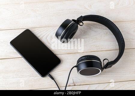 Smartphone und schwarze Kopfhörer auf einem Holztisch. Hören Sie Musik. Modernes, persönliches Gerät mit Touchscreen für Musik und Kommunikation. Draufsicht. Stockfoto