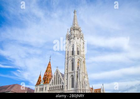 Der Turm der berühmten Matthias Kirche in Budapest, Ungarn. Die im Stil der Gotik erbaute Römisch-Katholische Kirche. Orangefarbenes Ziegeldach. Blauer Himmel und weiße Wolken darüber. Horizontales Foto. Stockfoto