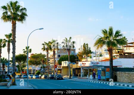 Paphos, ZYPERN - 13. FEBRUAR 2019: Stadtbild der Paphos Innenstadtstraße mit Palmen, Geschäften und geparkten Autos in Abendlicht Stockfoto