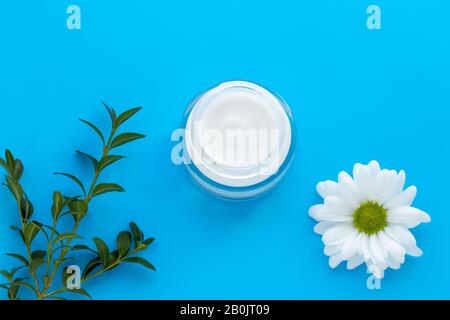 Glaskanne aus Gesichtscreme mit weißer Kamille auf blauem Hintergrund, Hautpflegekonzept. Kräuterlotion in der Flasche, natürliche Kosmetik mit Daisy Stockfoto