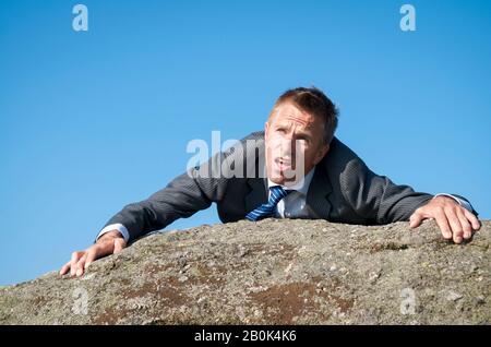Erschöpfter Geschäftsmann klammert sich an den Gipfel eines Felsenberges Stockfoto