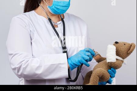 Weibliche Sanitäter halten braunen Teddybären mit Pfote in weißem Verband und hören Spielzeug mit Stethoskop, Konzept der Pädiatrie und Tierbehandlung Stockfoto