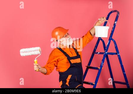 Arbeiten in der Werkstatt. Leitender Maler verwendet Rollen auf Leiter. Wand in Rosa malen. Professioneller Maler in Arbeitskleidung. Arbeiter malen Wand im Zimmer. Malerarbeiten mit Rolle für Männer. Stockfoto